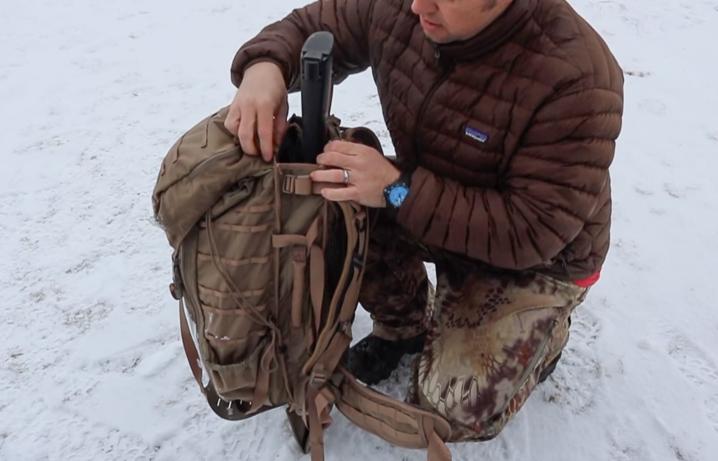 The author secures his Springfield Armory M1A SOCOM 16 rifle into the integrated rear scabbard of the Eberlestock Gunslinger 2 pack at the base of a snow covered mountain. The Eberlestock Gunslinger 2 review demonstrates how the rear scabbard system secures a full sized battle rifle for backcountry travel. The integrated rifle scabbard holds the SOCOM 16 tight against the pack body with minimal movement during climbing. Tactical packs built around rifle carry eliminate the need to hand carry firearms over demanding winter terrain. The Gunslinger II scabbard accommodates the M1A platform securely without requiring additional strapping or modification. Snow covered mountain terrain demands a hands free rifle carrying solution for safe and efficient movement. A properly secured rifle in a purpose built scabbard allows the shooter to focus on footing and navigation. The Gunslinger 2 Eberlestock was designed with the rifle as the central component of the entire pack system. Winter backcountry riflemen rely on integrated carry solutions to move farther and stay out longer in harsh conditions.
