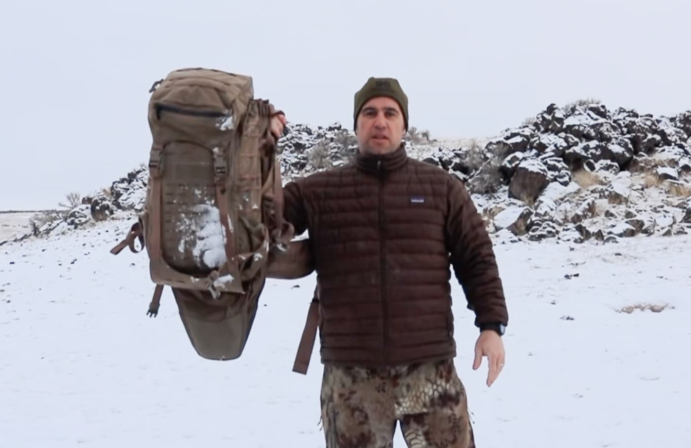 The author stands in a snowy outdoor setting demonstrating the key features of the Eberlestock Gunslinger 2 tactical pack. The Eberlestock Gunslinger 2 review covers the pack's integrated rifle scabbard, aluminum frame, and laser-cut MOLLE system. Tactical packs built for rifle carry require thoughtful construction to perform in harsh environments. The Gunslinger II features laser-cut Kevlar-reinforced MOLLE for strength without added bulk. An aluminum frame distributes weight efficiently across the back and hips during long carry distances. The SOCOM 16 rifle fits securely in the rear scabbard with minimal movement over uneven terrain. Ventilated back panels reduce heat buildup even during cold weather exertion. Exterior cinch straps allow snowshoes and additional gear to be secured outside the main compartment. A purpose-built tactical rifle pack integrates carrying, organization, and field utility into one cohesive system.