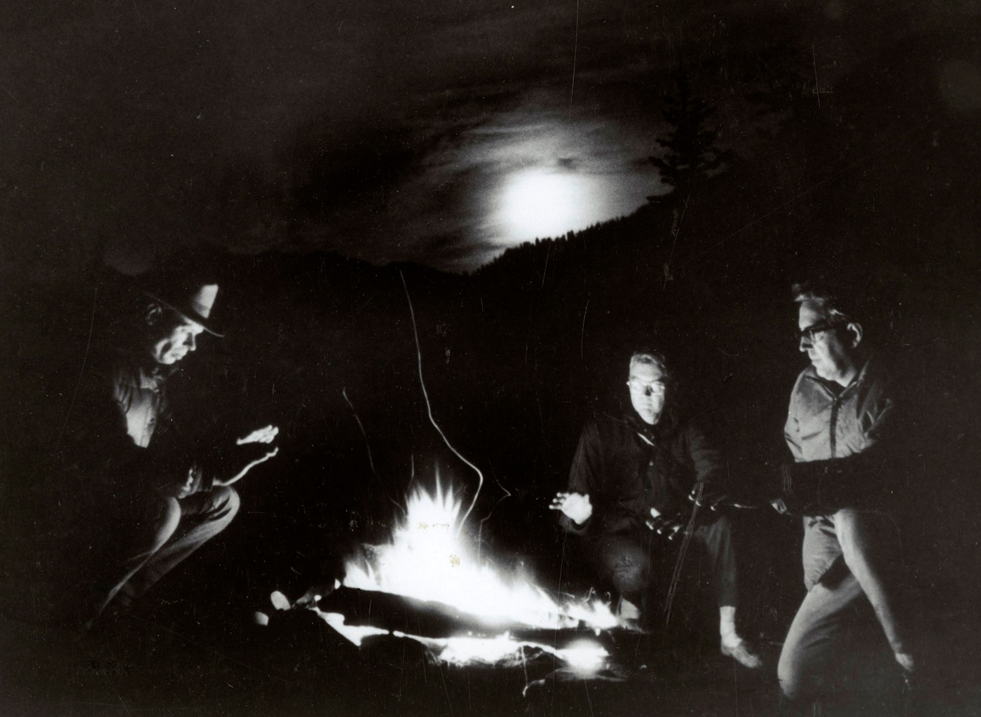 In this high contrast B&W photograph, we see three men around a campfire at night.