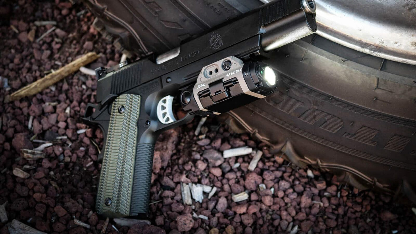 A Springfield Armory 1911-style pistol with a Viridian X5L light and laser unit mounted on the Picatinny rail below the barrel. The Viridian X5L review demonstrates practical home defense upgrades. Green laser technology improves target acquisition indoors. Combined light and laser units enhance low-light shooting capability. Non-standard response drills benefit from visible aiming references. Springfield Armory 1911 pistols accept standard rail-mounted accessories. Tactical lighting systems provide positive target identification. Home defense firearms require reliable illumination tools. The X5L offers dual-purpose defensive functionality.