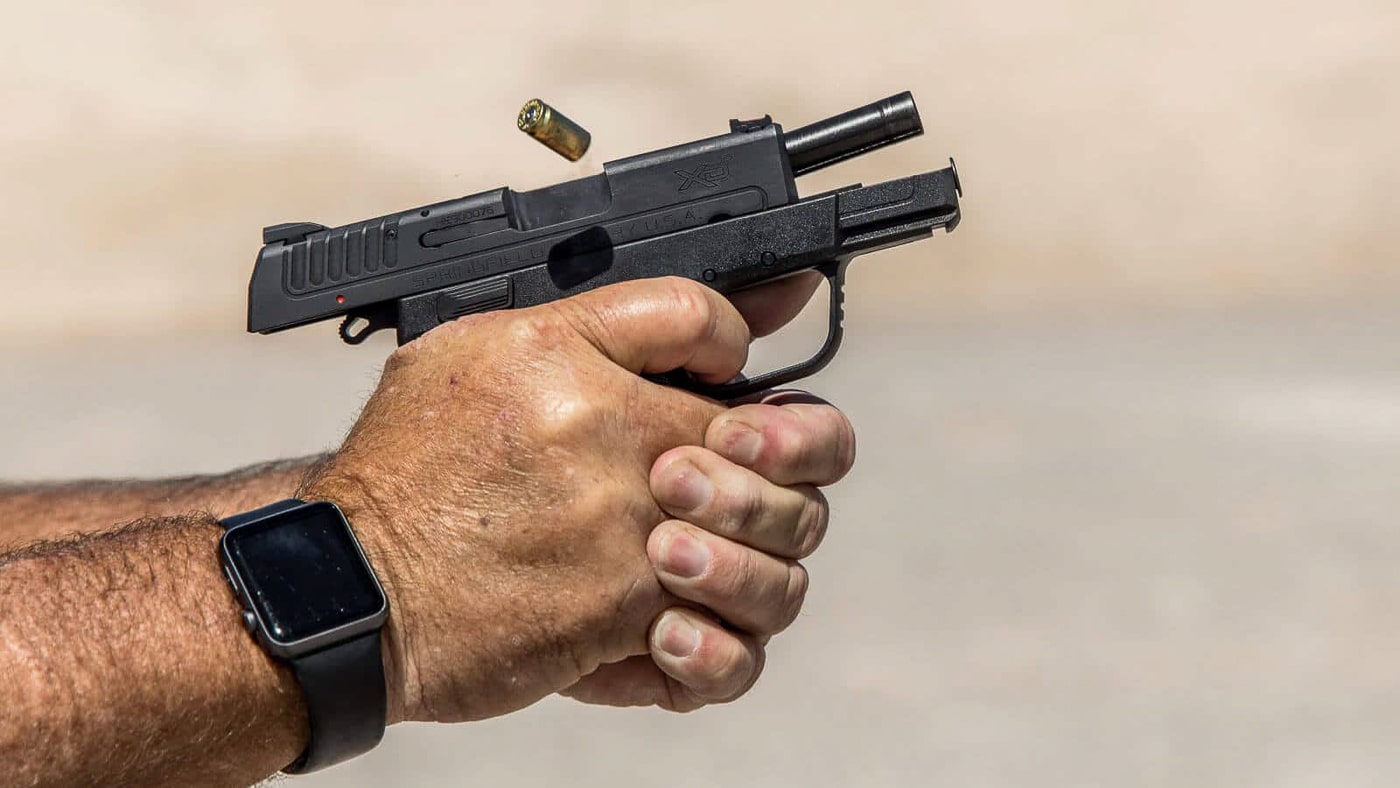 A close-up photograph shows a Springfield Armory XD-E pistol being fired at an outdoor shooting range with the slide in recoil position. The double strike capability of Springfield XD-E allows immediate follow-up hammer strikes on stubborn primers. DA/SA trigger systems provide mechanical redundancy for failure to fire situations. The Springfield Armory XD-E features a hammer-fired ignition system rather than striker-fired operation. Polymer-framed handguns can incorporate traditional double-action mechanisms for added reliability. Single-stack 9mm pistols offer excellent concealment while maintaining combat effectiveness. The XD-E decocker allows safe hammer lowering without trigger manipulation. Compact carry pistols benefit from deliberate double-action first pulls during holstering. Type one malfunctions resolve faster with second-strike hammer capability on centerfire cartridges.