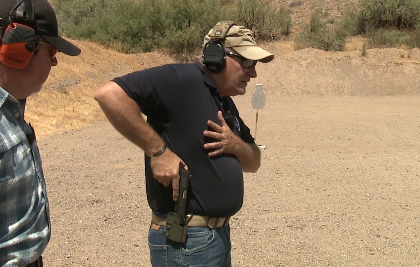 An instructor lifts a pistol vertically upward from a belt-mounted holster with the muzzle still oriented downward and the grip fully established. Pistol presentation requires vertical extraction movement. Drawing a handgun from a holster involves straight upward motion. Proper firearm clearance eliminates angular deviation. Handgun deployment maintains downrange safety during extraction. Defensive pistol techniques prioritize controlled lifting. Combat shooting mechanics use direct vertical paths. Holster retention systems release through upward pressure. Tactical handgun presentation avoids lateral muzzle sweep.