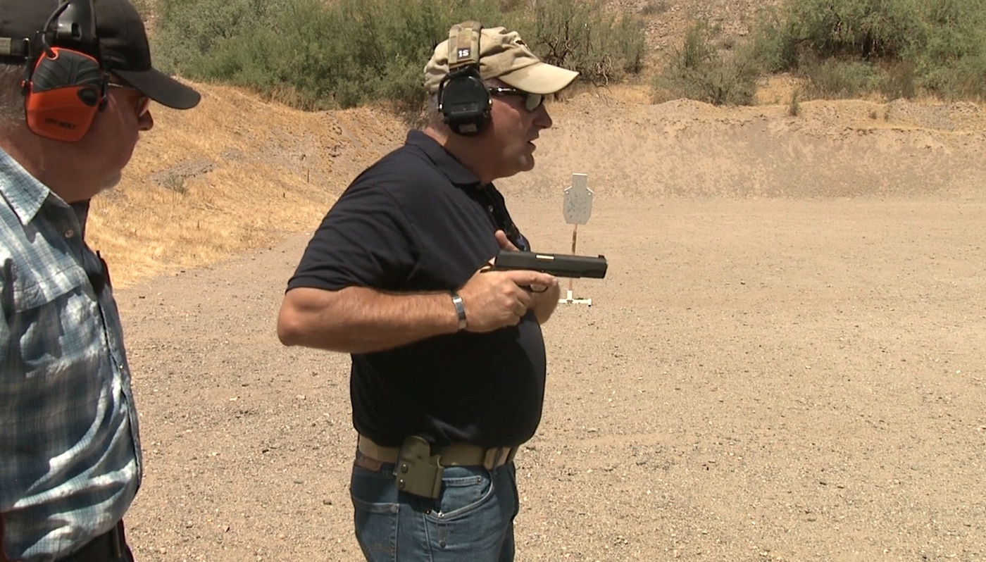 An instructor demonstrates the elbow drop phase with the firing arm moving downward and rearward while the pistol rotates forward toward the target plane. Pistol presentation involves controlled elbow positioning. Drawing a handgun from a holster requires deliberate arm mechanics. Proper firearm rotation eliminates lateral movement. Handgun deployment uses body-centered motion paths. Defensive pistol techniques emphasize elbow-driven rotation. Combat shooting mechanics minimize upper body twisting. Holster clearance transitions into forward alignment. Tactical handgun presentation integrates natural body positioning.