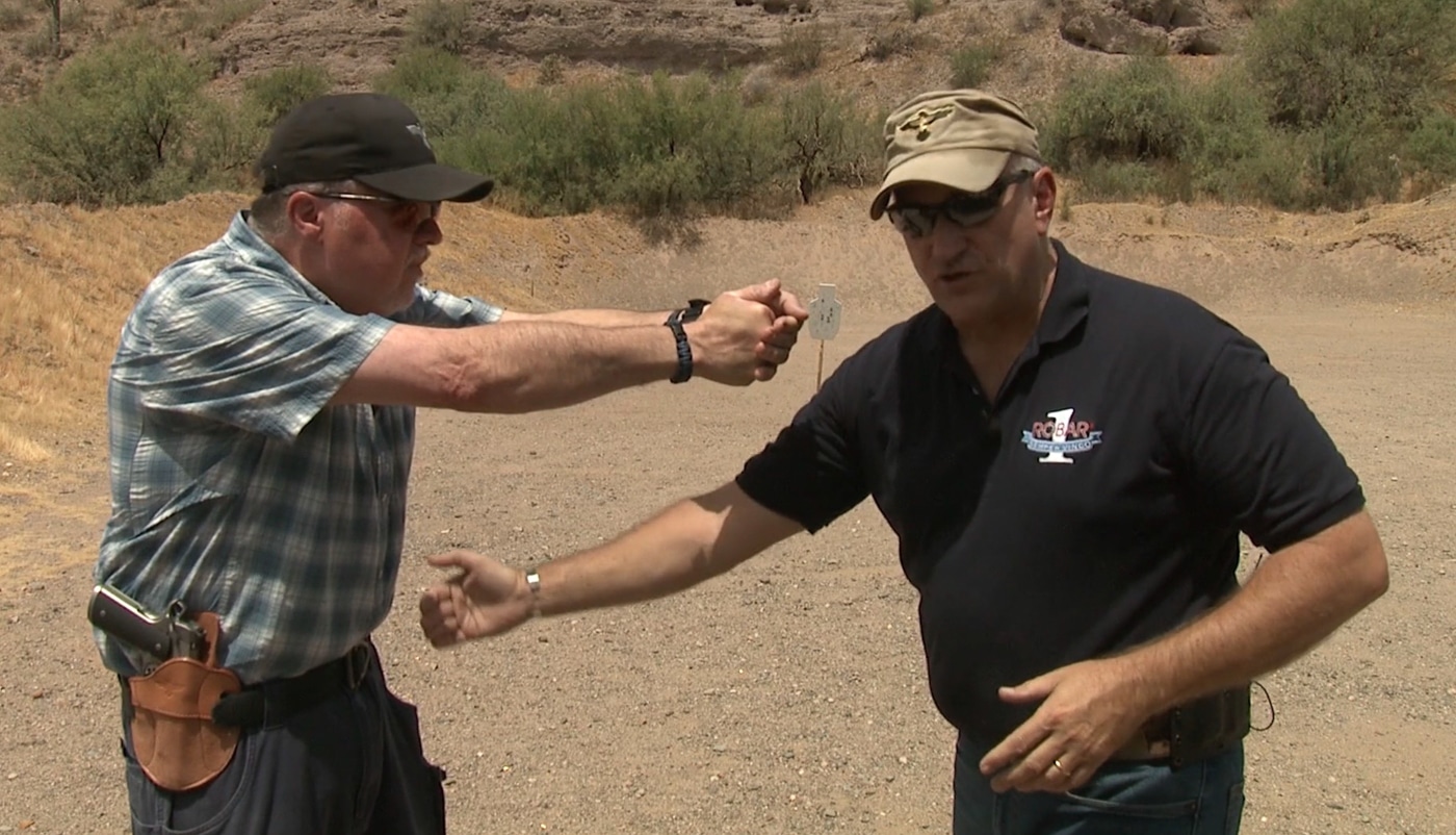 An instructor places their hand on a student's back to guide proper forward lean, ensuring the chest positions correctly over the belt buckle during stance instruction. Learning a shooting stance requires correct weight distribution. Firearms instruction on shooting stances emphasizes forward positioning. Chest placement affects recoil absorption. Shooters control muzzle rise better. Forward lean prevents backward rocking. Upper body mechanics improve accuracy. Instructors provide hands-on corrections. Proper posture enables faster shooting.