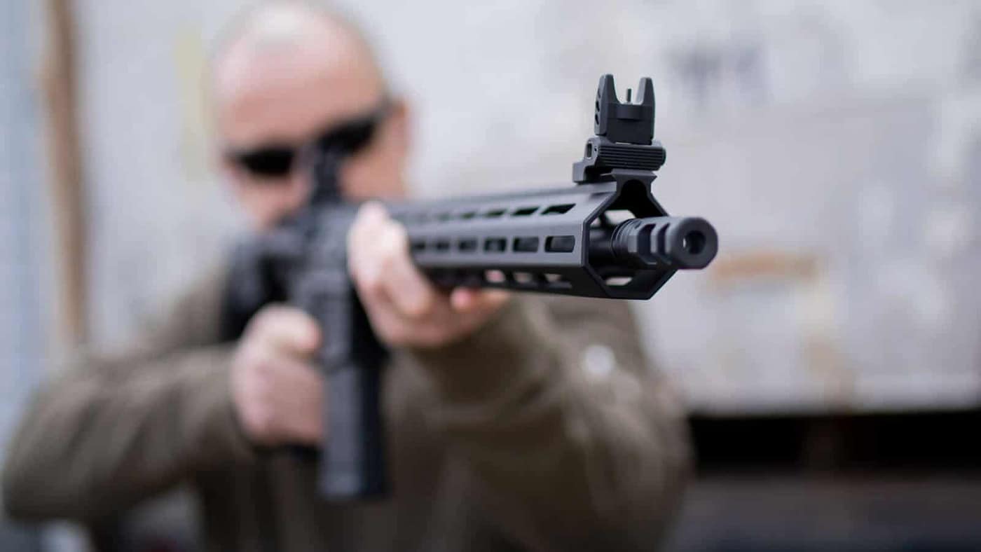 A shooter practices rapid fire with a rifle at an outdoor shooting range, maintaining an aggressive stance while engaging a close-range target. Non-standard response drills simulate life-threatening encounters. Defensive rifle training emphasizes speed over precision. Close-quarters shooting requires immediate incapacitation capability. Tactical response techniques address edged weapon threats. Rifle training drills build combat-ready fundamentals. Center mass engagement ensures maximum stopping power. Fighting stance principles support recoil management. Defensive shooting courses prepare for violent encounters.