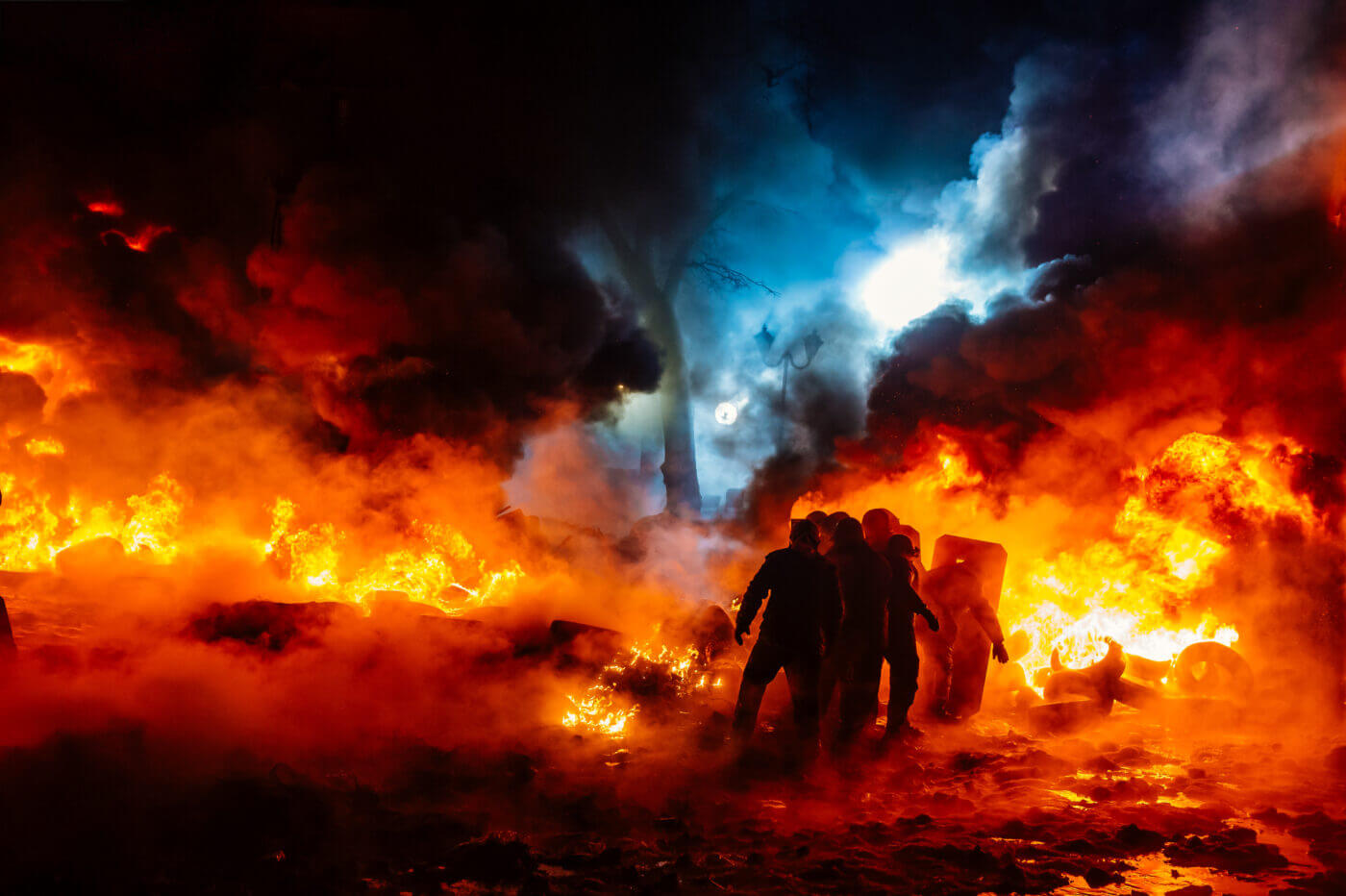 A group of wilderness firefighters in full protective gear battles a massive wildfire approaching the edge of a densely populated urban area. Thick smoke rises into the sky, partially obscuring the outline of the city skyline in the background. Firefighters wield hoses and tools to control the advancing flames, while others monitor the fire's spread in the dry, brush-filled landscape. This image highlights the vulnerability of urban areas to natural disasters like wildfires and underscores the importance of emergency preparedness and situational awareness. The scene is a stark reminder of the risks posed by extreme weather events, particularly in areas where rural landscapes meet urban environments. The firefighters’ efforts represent a crucial line of defense for the community, infrastructure, and utilities threatened by the fire.