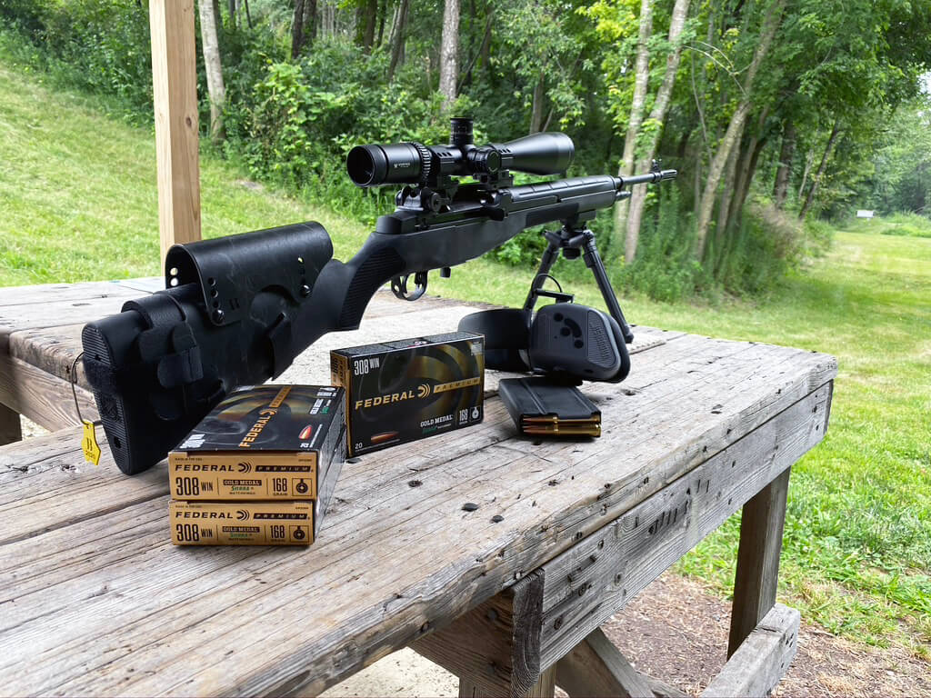 In this digital photograph, the author has installed the Bradley cheek rest on her M1A rifle. She is at a private shooting range with the gun attached to a bipod and resting on a shooting bench. Visible in the photo are .308 Winchester cartridges which are compatible with the 7.62x51mm and will fire in the M1A. Also visible is a metal box semi-automatic rifle magazine and an electronic hearing protection device.
