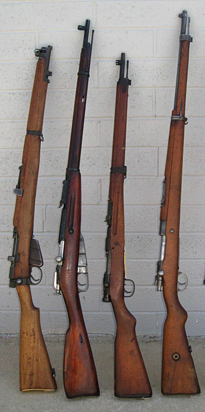 Four vintage military rifles, including the British No. 1 Enfield, Russian Mosin-Nagant, Japanese Arisaka, and German K98 Mauser, are displayed leaning against a wall at a Vintage Military Rifle Games competition. The rifles' wooden stocks and metal finishes highlight their historical craftsmanship and military significance. This lineup reflects the diverse firearm designs used by nations during the early 20th century. The image captures the spirit of the Vintage Military Rifle Games, celebrating history and marksmanship with these iconic rifles.