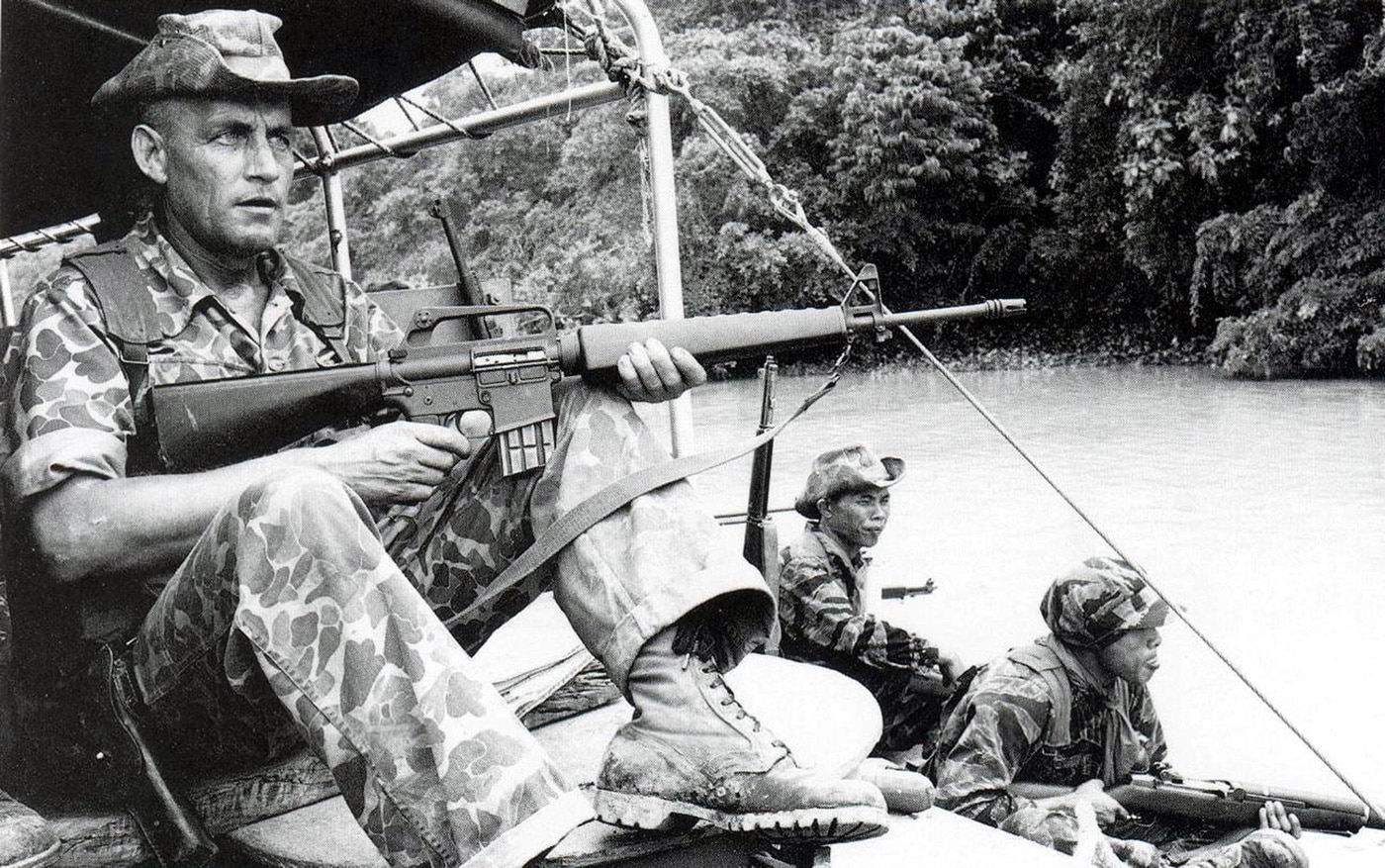 A U.S. Army advisor armed with an AR15 Model 602 rifle carbine.  Note the absence of a forward assist device and that the ARVN soldiers he is embedded with are armed with M1 Garand rifles. Tiger stripe camo and jungle camo us soldiers.