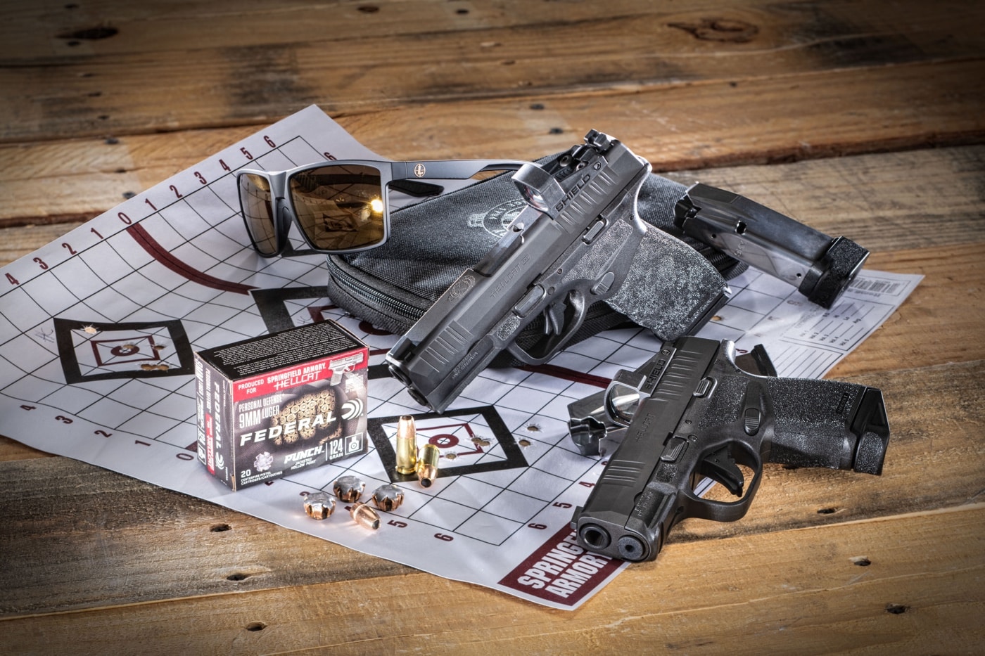 In this photo, the author of the article has arranged a Springfield Hellcat and Hellcat Pro with a scored target, ammunition, a soft sided care, a gun magazine and eye protection glasses that look like sunglasses.