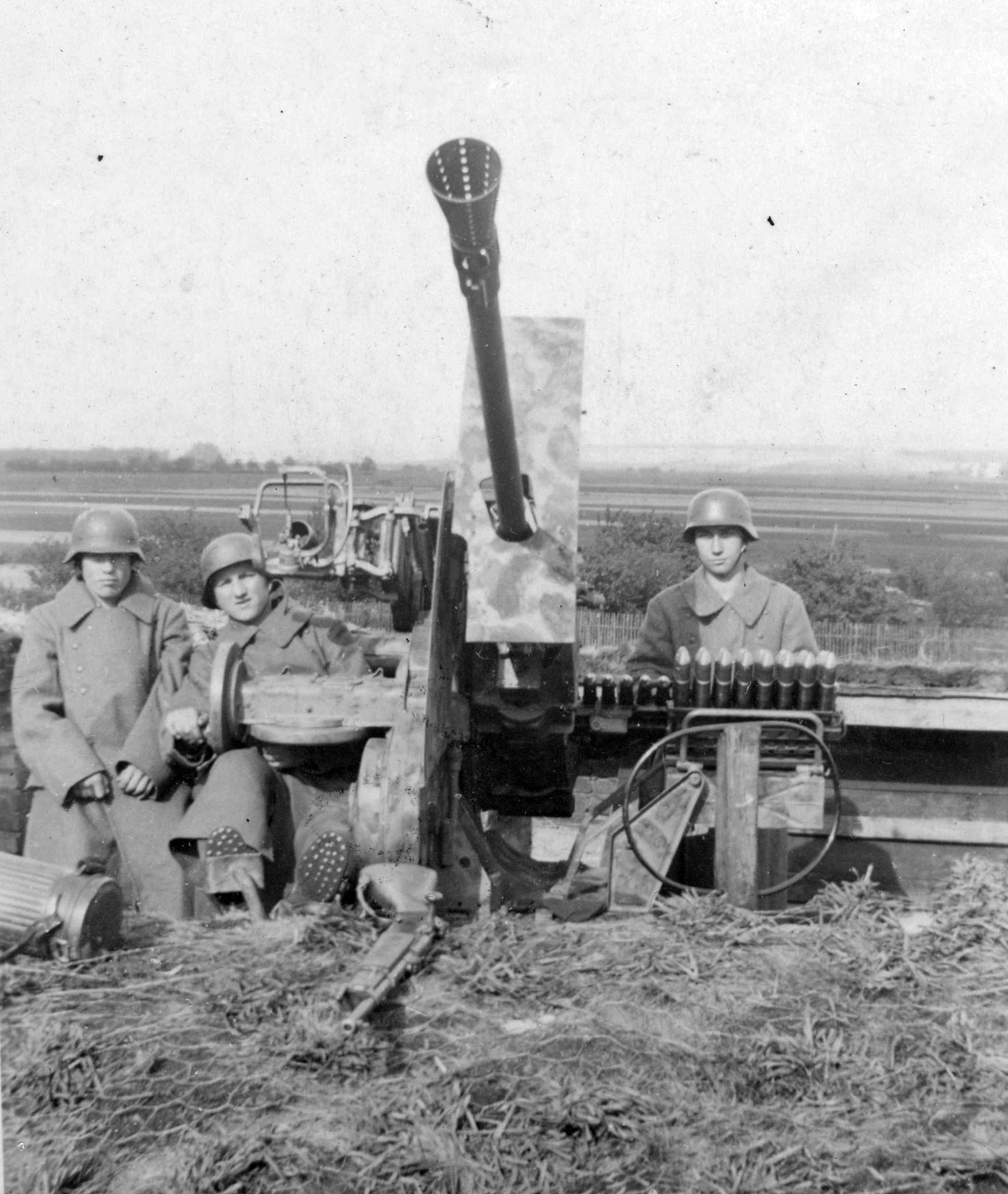 This photo shows a German 37mm AA gun set up to defend the Ploesti oil facilities in Romania. The image shows three German soldiers with ammunition and the gun in a trench. In the foreground is a blot action rifle. The soldiers are all wearing helmets and uniform trench coats. One of the soldiers has visible hobnailed boots. 