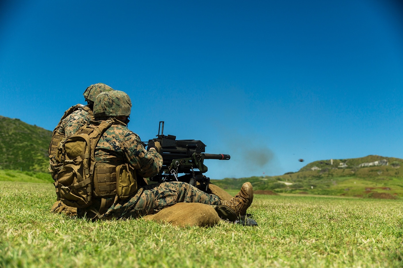 Shown is a pair of Marines shooting a Mk-19 grenade launcher during training in Japan. They are wearing their green camo utilities and helmets with full body armor. There is a hill in the background, green grass everywhere and a bright blue sky.