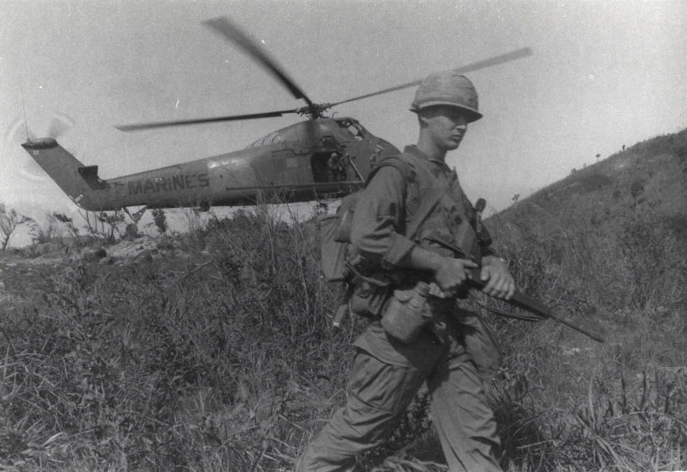 In this image, we see a Marine equipped with a M16 rifle, helmet, canteen, backpack and utility uniform walking down a grassy hill and away from the helicopter that transported him there. The image highlights the helicopter role in modern warfare — something that started in the Vietnam era circa 1962. Choctaws like this one had tail-dragger rear fuselage and landing gear. The utility helicopter was adopted by many allied militaries around the world including the French Air Force and German Air Force.