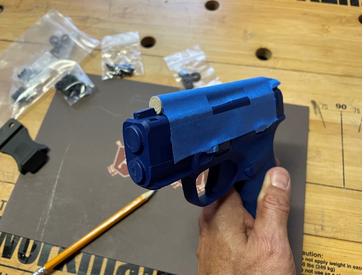 In this photo, the author shows how he used painters blue tape to attach a wooden dowel to the top of the Hellcat blue gun. When the Kydex is folded over the top of the gun, the dowel will ensure that there will be room to spare for the front sight to enter and exit the rig smoothly. With the dowel in place, you can use a heat gun to warm the Kydex to achieve a precise fit. 