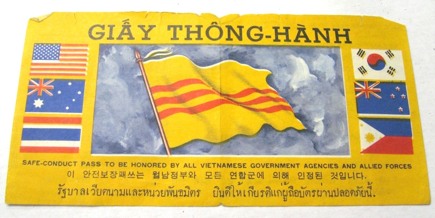 A rare safe conduct pass is shown in this photo. The obverse of the paper pass has a large South Vietnam flag in the center and is flanked by smaller flags of the United States of America, Australia, Thailand, South Korea, New Zealand, and the Philippines. The reverse has an official stamp, is signed, and contains Vietnamese text explaining the assistance they will receive if they surrender. Both sides contain English text reading "Safe-conduct pass to be honored by all Vietnamese government agencis and allied forces." The obverse contains a similar phrase in both Thai and Korean.