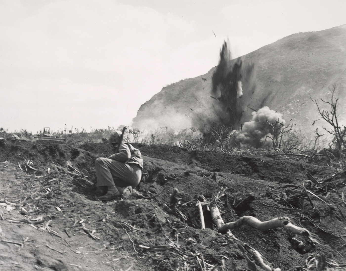 We see naval gunfire hitting Japanese positions at the base of the volcano. In the foreground is a U.S. Marine waiting for the firing to lift so he can move forward.