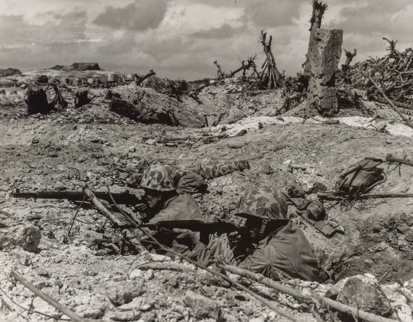 In this photo, two Marine infantrymen stay low in their trench while U.S. Navy ships continue the bombardment of the volcano island. Part of the United States Armed Forces amphibious warfare strategy, naval warfare typically included the use of battleship artillery hitting the blockhouse and pillboxes used by the Imperial Japanese Army.