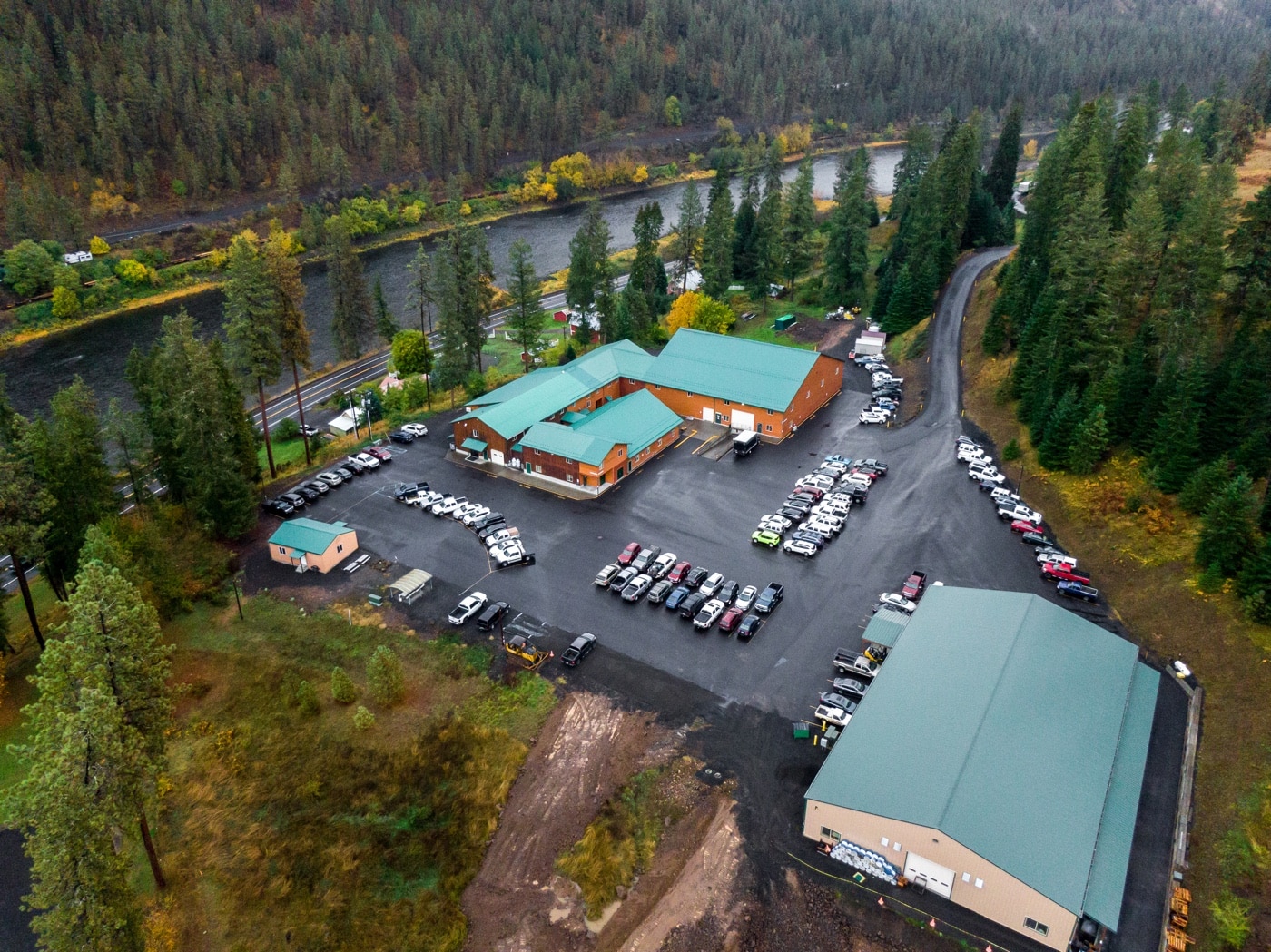 In this digital image, we see the Nightforce Optics manufacturing facility from the air. The facility features two main buildings with a smaller auxiliary building. There is a large parking lot with many cars and trucks parked in it. There are many motor vehicles here. In the surrounding area we can see a river, mountains and evergreen trees.