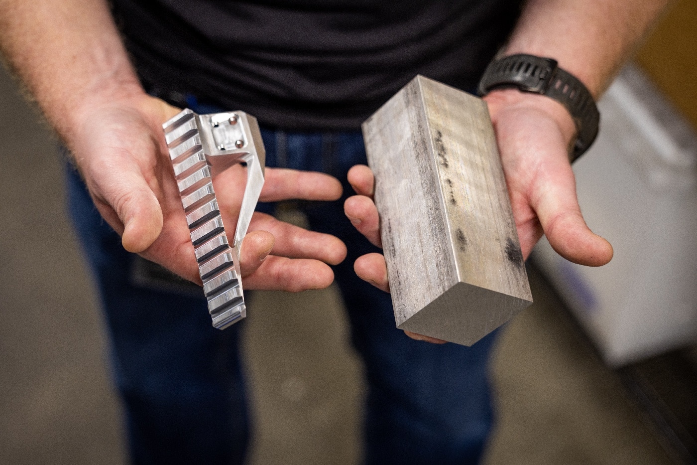 In this photo, a machinist holds an unworked piece of aluminum in one hand with a finished scope mount in his other hand.