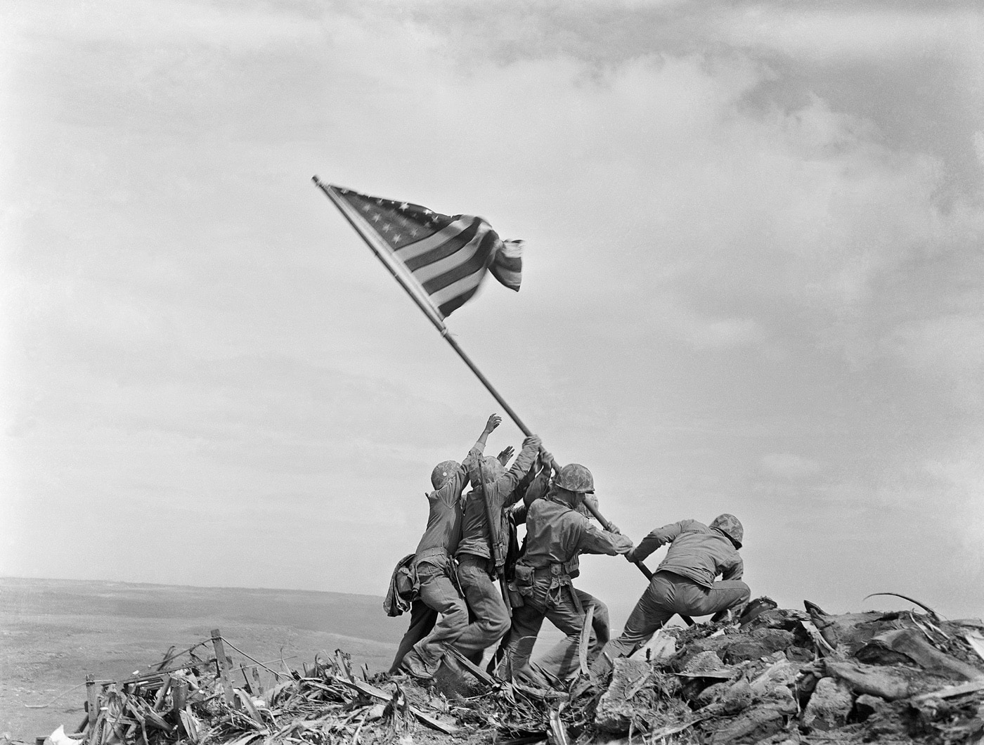 In this photograph, Associated Press photographer Joe Rosenthal took a film photograph of 4 U.S. Marines raising the flag of the United States of America on Mount Suribachi during the Battle of Iwo Jima in WW2. Part of the Pacific War, Iwo Jima was a volcanic island that had to be taken to provide P-51 Mustang escort fighter aircraft to Boeing B-29 Superfortress and Boeing B-17 Flying Fortress air raids on Japan. This helped to lead to the surrender of Japan and the Victory over Japan Day (VJ Day). 