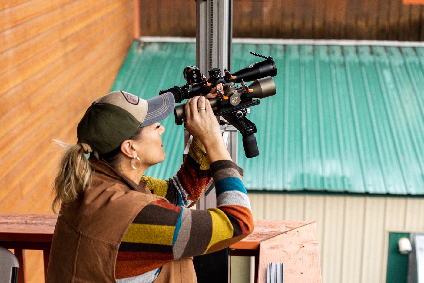 In this photograph, a woman employed by Nightforce Optics performs testing on the scopes. She is wearing a hat and is outside on a second story balcony. A green metal roof is visible in the background.