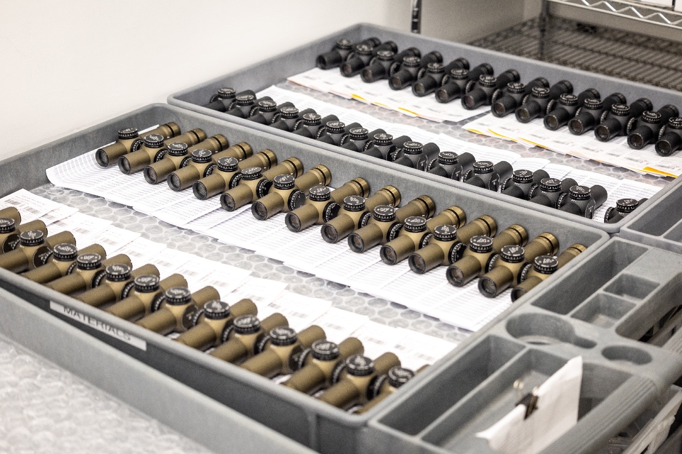 Two trays of Nightforce Optics scopes are shown in this photo. The closest tray has anodized scopes in a flat dark earth color. The farther tray is full of scopes with a black anodized finish.