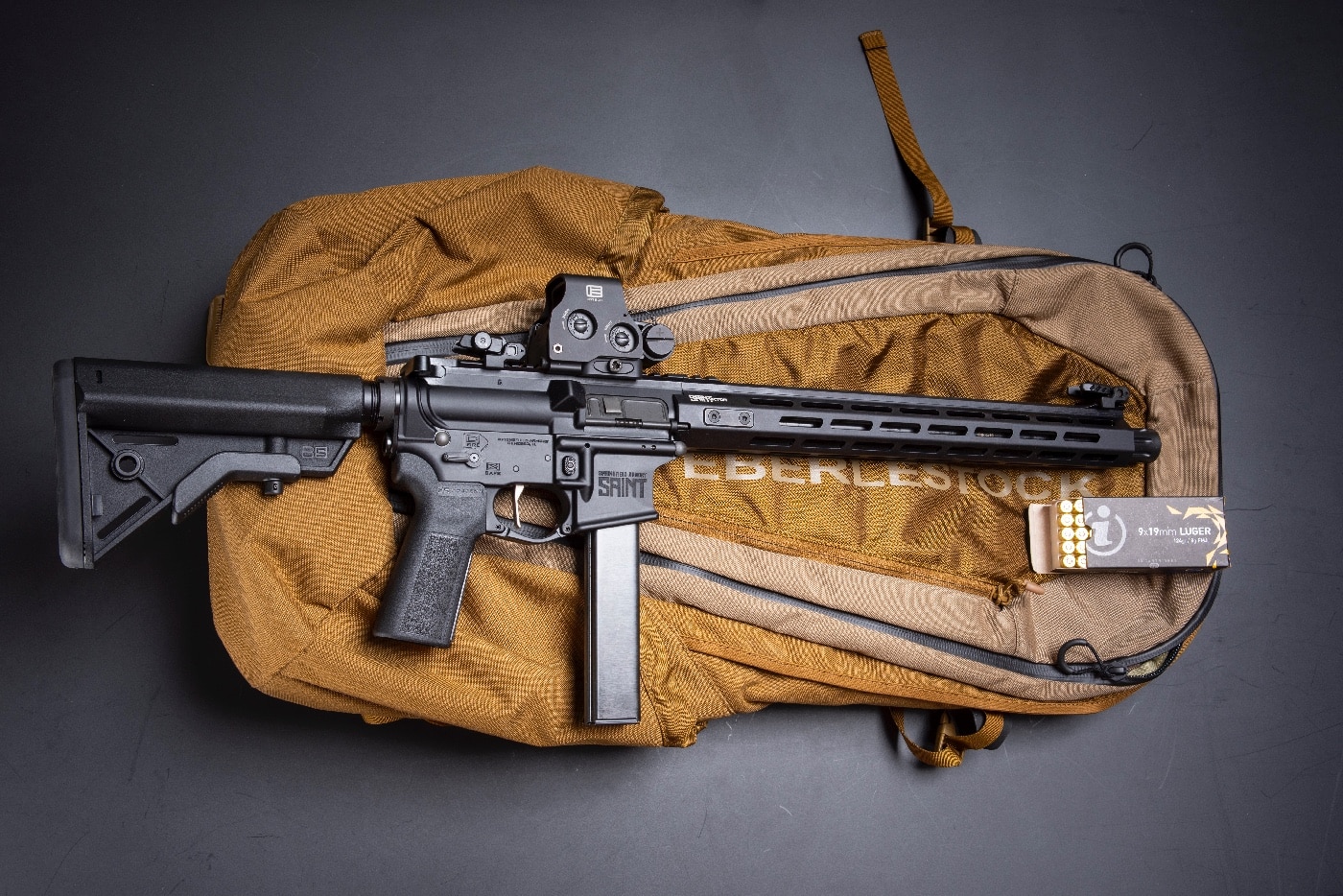 In this photo, a 9mm ar-15 is shown along with a carrying case and ammunition for a trip to the shooting range for target practice. Some people buy a pistol version of an AR and add a pistol brace to it. While that is a perfectly legitimate option, a standard rifle length carbine takes your ammunition and gives it extra velocity and energy which may prove helpful in a defensive shooting. Plus, a rifle stock increases your points of contact on the gun for improved accuracy.