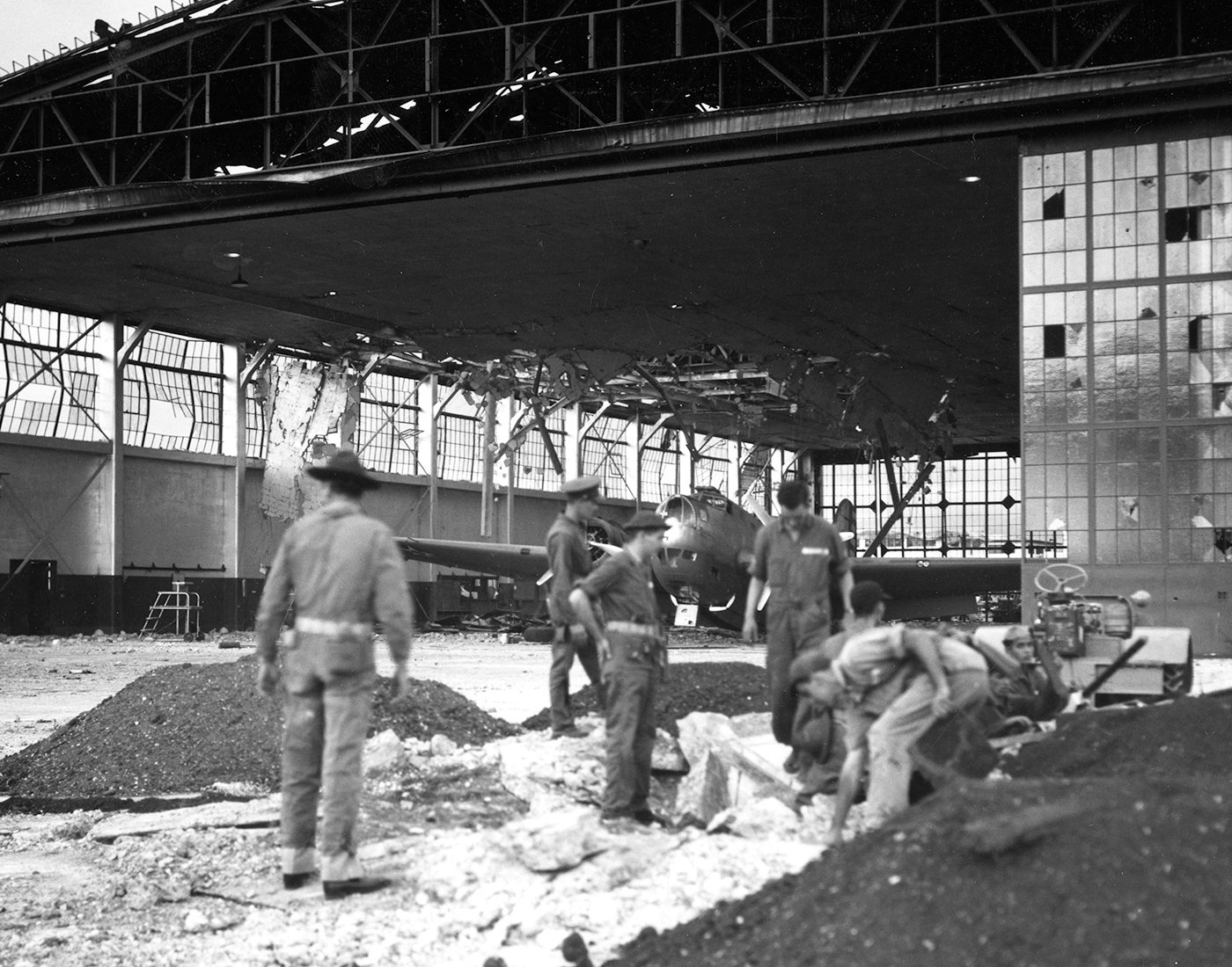 This photo, taken on the day of the Pearl Harbor attack, shows a damaged B-18 Bolo in a hanger. The hangar is damaged. Soldiers in the foreground are setting up a crew served machine gun in a crater left by a Japanese bomb.