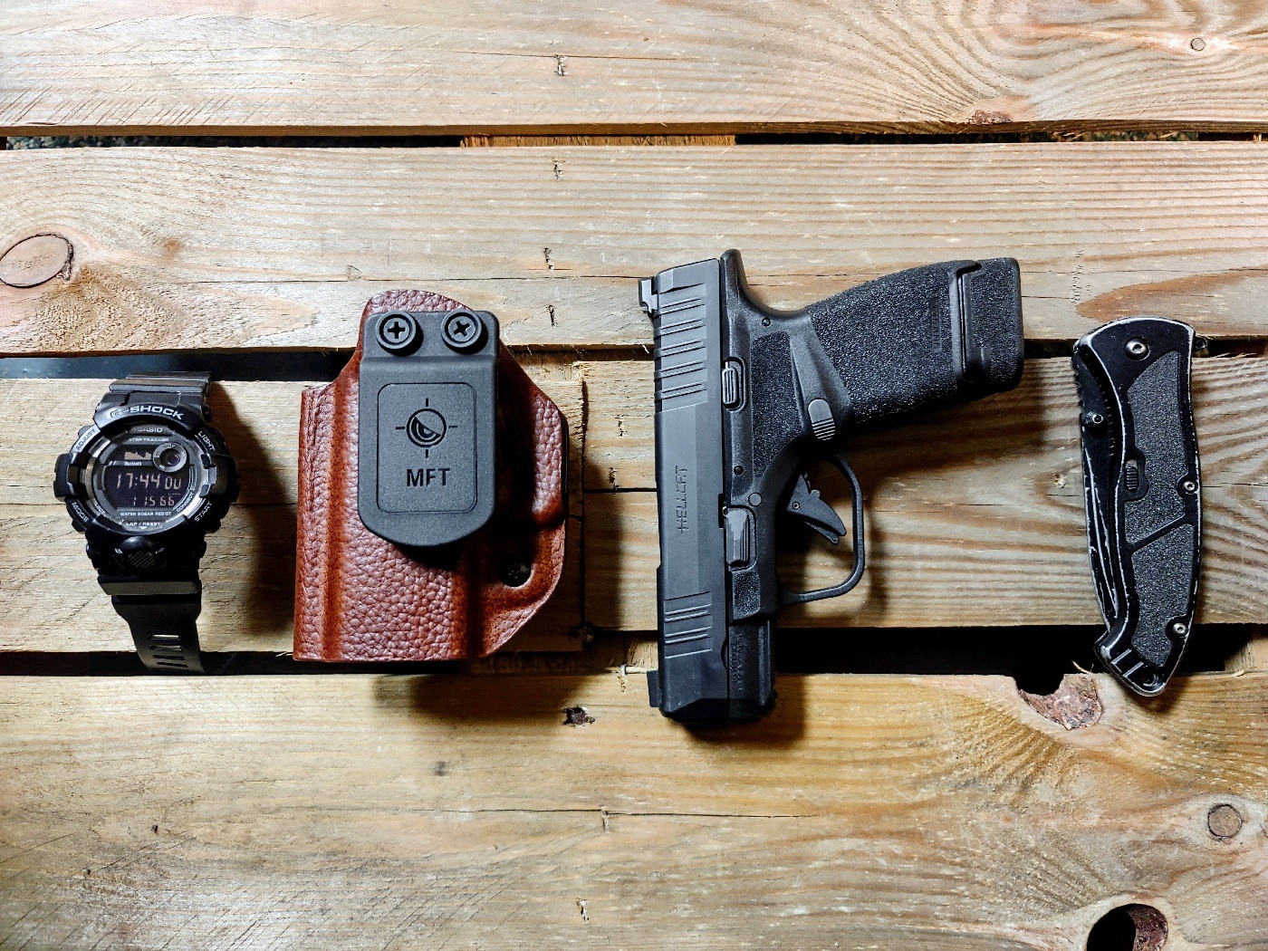 The photo shows a MFT holster along with a digital watch, a folding knife and a Springfield Armory Hellcat.