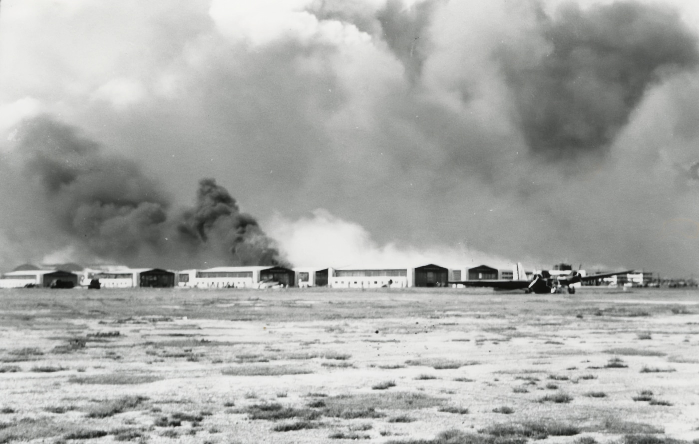 In this photo, we see a damaged B-18 Bolo in the foreground with smoke and fires burning in the distance. The bomber was hit while on the ground at Hickam Field. The black smoke in the background is from the U.S.S. Arizona, a battleship that was destroyed during the sneak Attack on Pearl Harbor. The attack on Pearl Harbor was a surprise military strike by the Imperial Japanese Navy Air Service upon the United States against the American naval base at Pearl Harbor in Honolulu, Hawaii, just before 8:00 a.m. on Sunday, December 7, 1941.