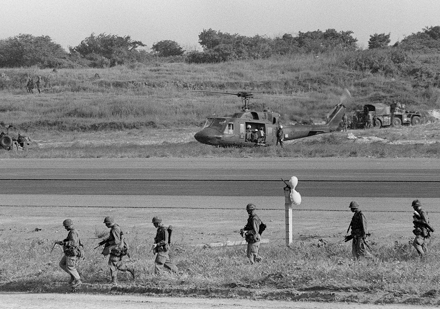 In this photo, we see Marines engaged in Operation Urgent Fury advance along a road on Grenada. A UH-1 Iroquois helicopter prepares to take off to support their mission. 