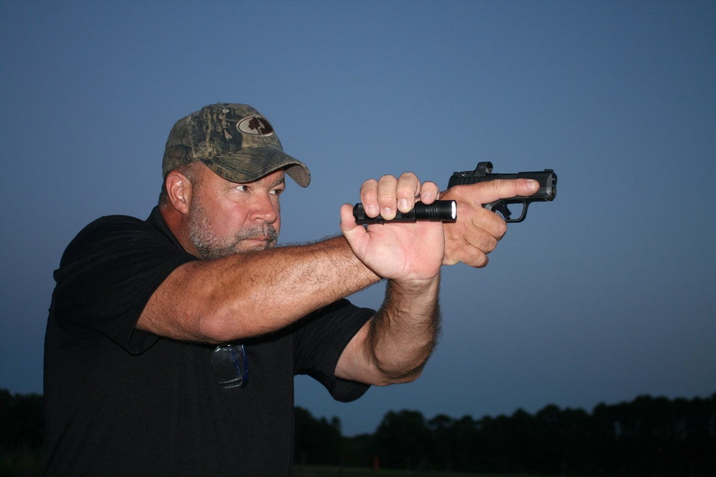 Photo of a shooter practicing in a shaded outdoor area, micro pistol drawn and light activated. The illumination casts a strong cone forward, clearly lighting up the cardboard silhouette target. The shooter is braced in a ready stance, using both hands to control the gun. Dusk conditions offer just enough ambient light to simulate real-world threat environments. This demonstrates a practical application of using lights for micro-compact pistols where threat ID and proper illumination matter.