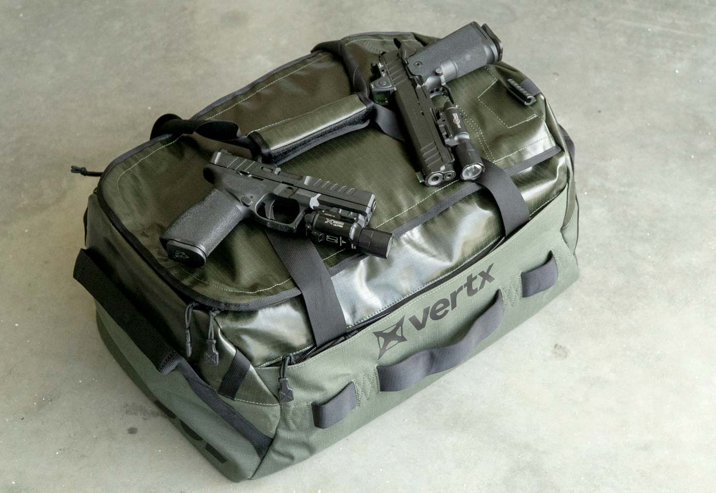The Vertx RLT 50L Duffel Bag shown closed with two Springfield Armory pistols placed on top for size comparison. The tactical duffel's compact exterior dimensions are demonstrated with the firearms providing visual scale reference, highlighting the bag's efficient design.