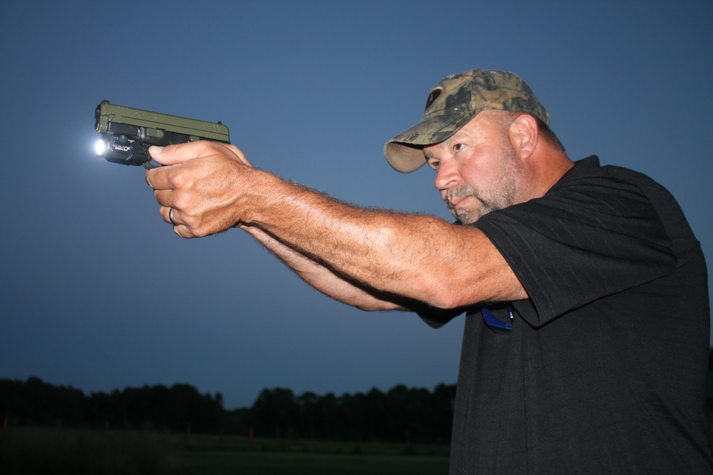 The author is captured during live fire at dusk, holding a micro-compact pistol in a two-handed firing grip. The weapon-mounted light is on, projecting a tight beam toward the target area. The fading daylight creates shadows behind the shooter while the light ensures clear identification downrange. This setup reflects how using lights for micro-compact pistols supports responsible and fast engagement in realistic low-light environments. The photo highlights practical training, not staged drills.