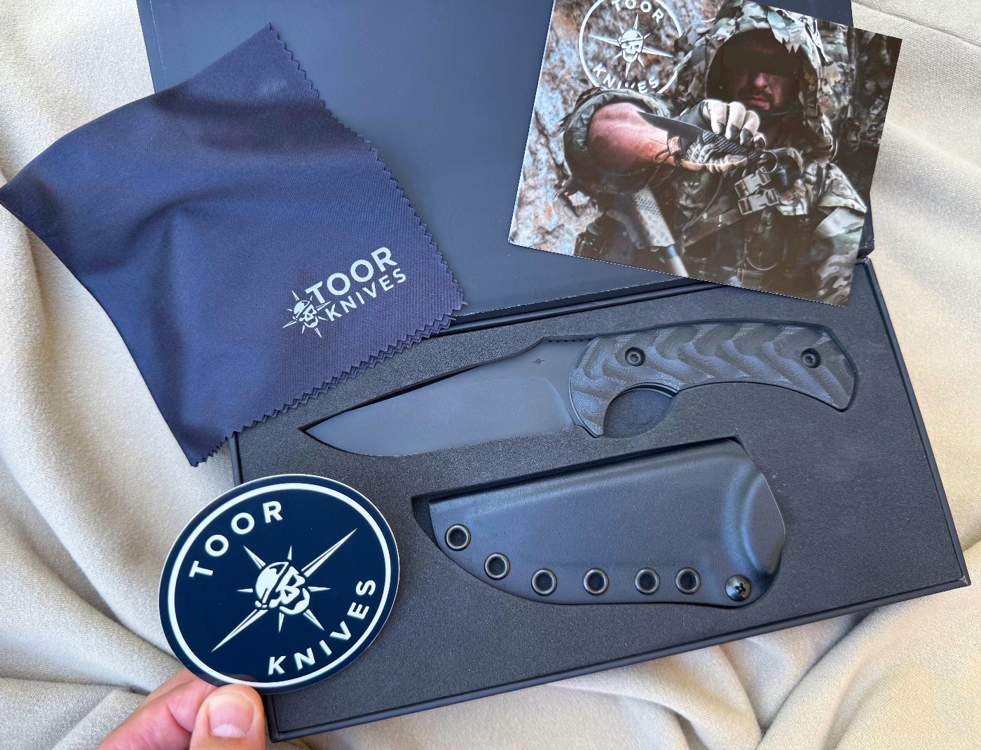 Photo showing the complete Toor Knives Mullet S kit including the knife, black Kydex sheath, FrogLube lubricant, welcome card, Toor sticker, and microfiber cloth. Each item is neatly arranged inside a black presentation box with magnetic closures and custom foam inserts. The setup reflects the premium nature of the product and its American manufacturing quality. The Mullet is a fixed blade knife designed for tactical and everyday use. The packaging reflects a high-end approach common with U.S.-made knives at this price point.