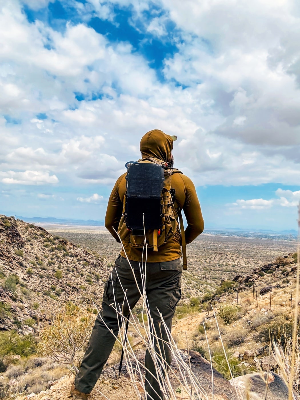 Dark Energy review shows the author hiking up a rocky desert incline with the Spectre solar panel attached to his backpack harness. The ultralight panel’s hex-patterned CIGS cells face the bright sun, maximizing off-grid power collection. A braided charging cable disappears into his pack, indicating continuous energy transfer to a battery inside. You can see rugged pack straps and reinforced edges built for field use. This image demonstrates portable solar charging effectiveness in real-world backcountry conditions. It underscores reliable power generation while on the move.