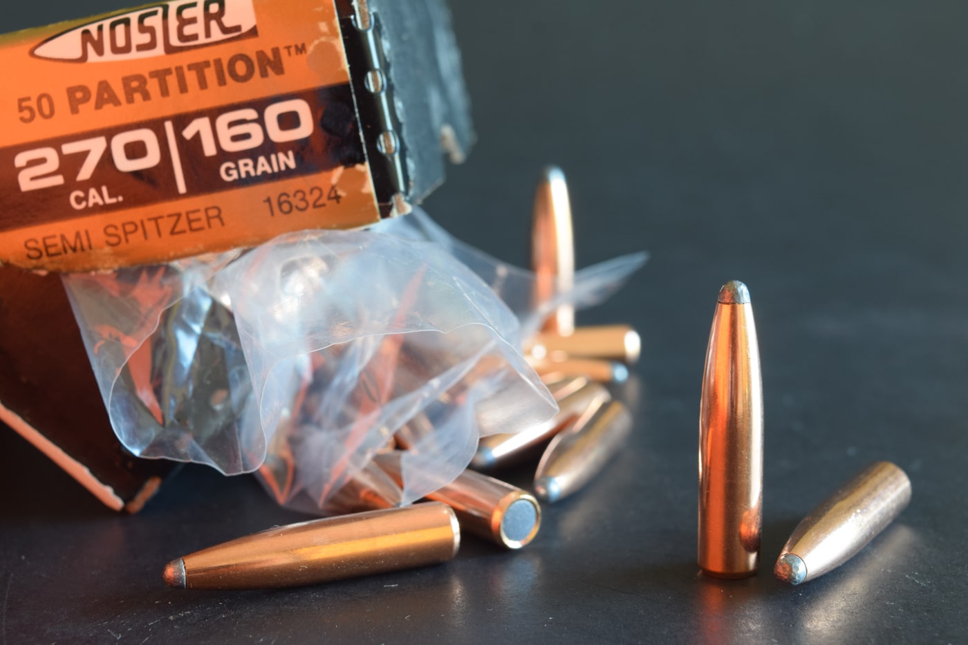Macro shot of three Nosler Partition spitzer bullets (160-grain) for the .270 Winchester displayed on a weathered wooden bench. The copper-jacketed boattail projectiles feature the Partition core designed for controlled expansion on medium and big game hunts. Their sleek spitzer profile promises high ballistic coefficient and flat trajectory beyond 300 yards. At 160 grains, these bullets deliver deep penetration with high sectional density for clean energy transfer on deer or elk. Spiral machining lines circle the jacket above the boat-tail, reflecting rifling twist rates common to .270 Winchester rifles. These components underscore the .270 Winchester’s legacy as America’s deer cartridge, hailing from its necked-down .30-06 origins. The image highlights the precision craftsmanship and hunting-grade performance associated with modern .270 Winchester loads.