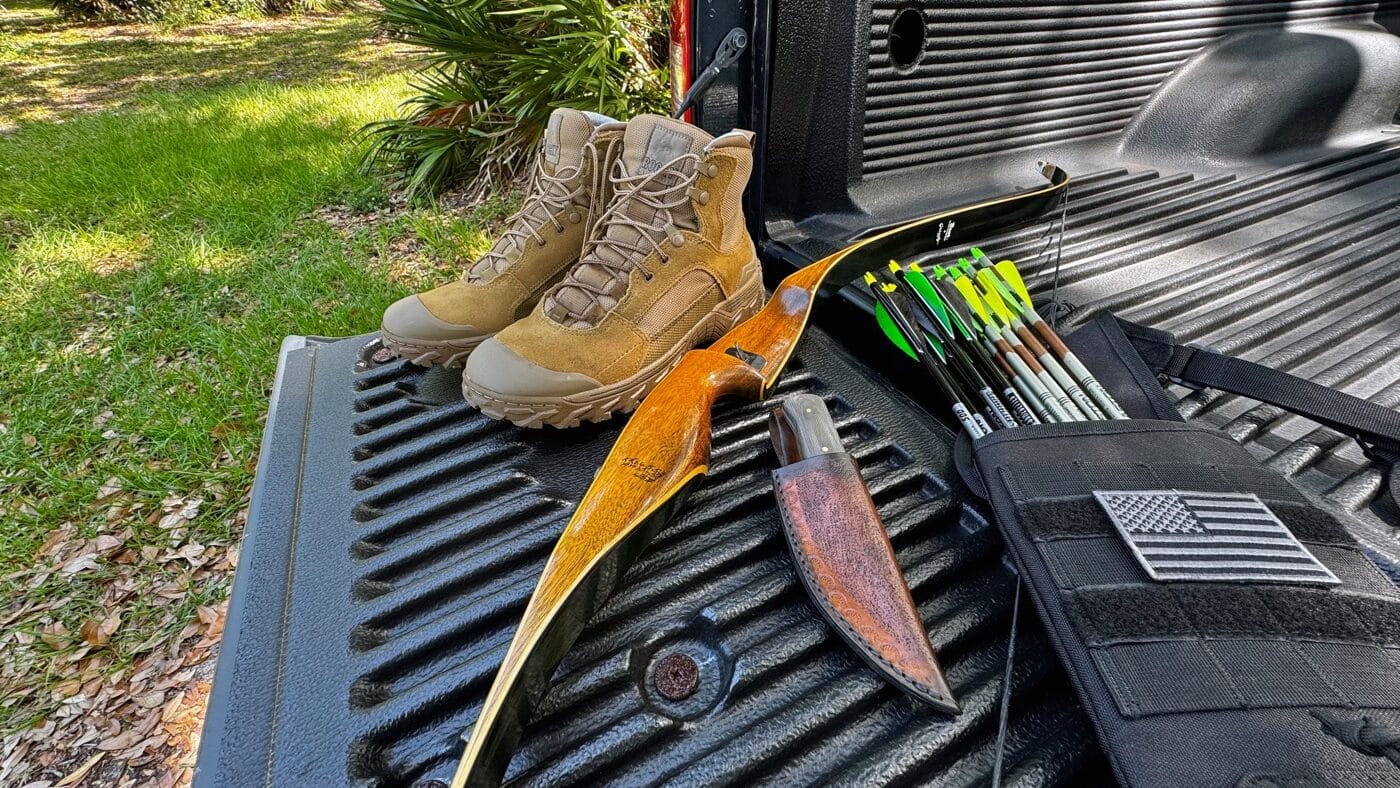 Pair of Rocky Mountain Combat Boots placed beside a recurve bow, arrow quiver, and loose arrows on grassy ground. The coyote tan boots show light dusting and creasing from test hikes. The bow’s wood finish and string are clearly visible, tying into an outdoor or shooting scenario. Fallen leaves and grass blades frame the boots, suggesting a natural setting. This photograph complements the Rocky Mountain Combat Boot Review by illustrating multi-sport functionality.