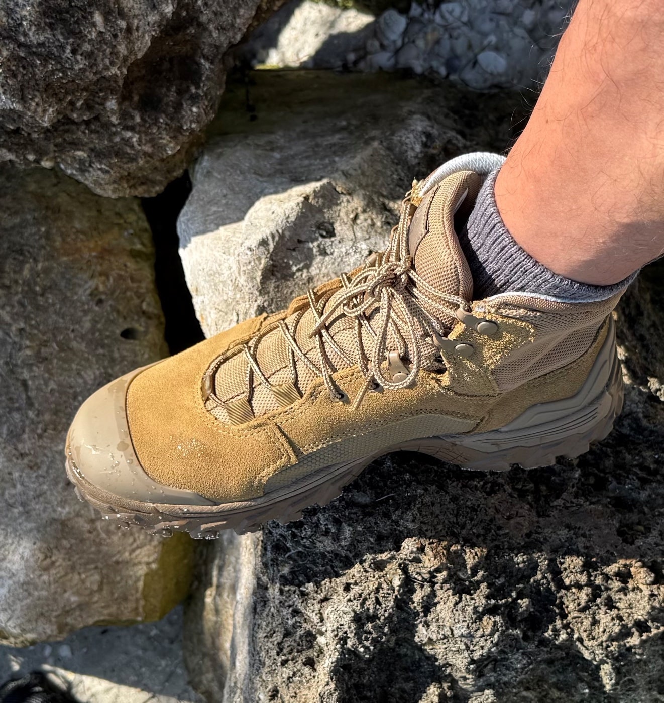Rocky Mountain Combat Boots walking across a rocky causeway streaked with algae and shallow water. The coyote tan leather contrasts with the darker, slippery surface of the wet stones. Outsole lugs are dug into the jagged terrain, demonstrating grip in challenging conditions. Small bits of sand cling to the midsole edge and the lower leather, indicating recent traversal through loose terrain. This image belongs to the Rocky Mountain Combat Boot Review and emphasizes traction on uneven, wet surfaces.