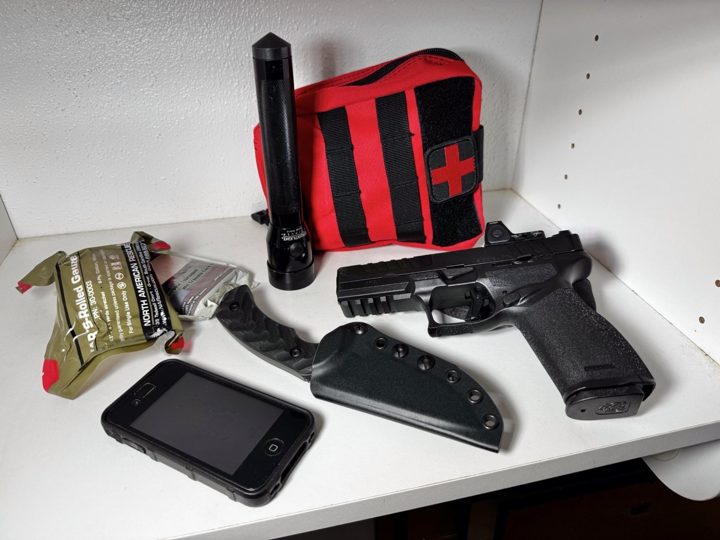 The photo features a compact defensive pistol positioned next to a collection of first aid gear, including trauma medical supplies such as a tourniquet, pressure bandages, gauze, and possibly a chest seal pack. This arrangement highlights a comprehensive approach to home defense, where being ready to treat injuries is just as critical as having tools for protection. In the event of a home invasion, wounds can happen quickly, and immediate access to trauma gear can mean the difference between life and death before emergency responders arrive. Storing medical supplies together with your defensive firearm at a designated rally point is a practical strategy for how to survive a home invasion. The combination allows for immediate security and medical intervention in chaotic situations. Familiarity with both emergency medical care and firearm use is an important part of home safety planning. Including both elements ensures your household is better prepared to deal with violence, accidents, and other unpredictable outcomes.