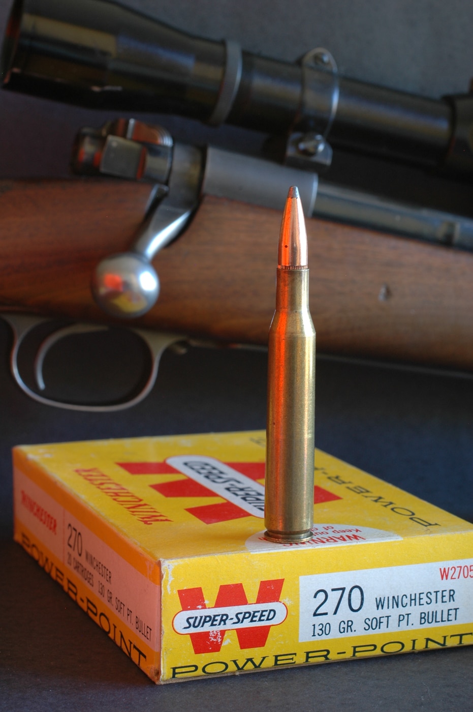 A .270 Winchester cartridge poised atop a vintage Winchester box of 130-grain Power-Point hunting ammunition. The aged cardboard display box shows the classic red-and-gold logo and faded text. The .270 round’s brass case and copper-alloy jacket glint in soft ambient light. The tapered shoulder and long neck of the case emphasize the cartridge’s design lineage from the .30-06 Springfield. The bullet’s sleek ogive hints at its high ballistic coefficient and supersonic trajectory potential. The packaging’s worn edges and scuffed corners evoke decades of gun cabinet history. This image highlights the cartridge’s reputation for flat-shooting performance and mild recoil on medium-game hunts.