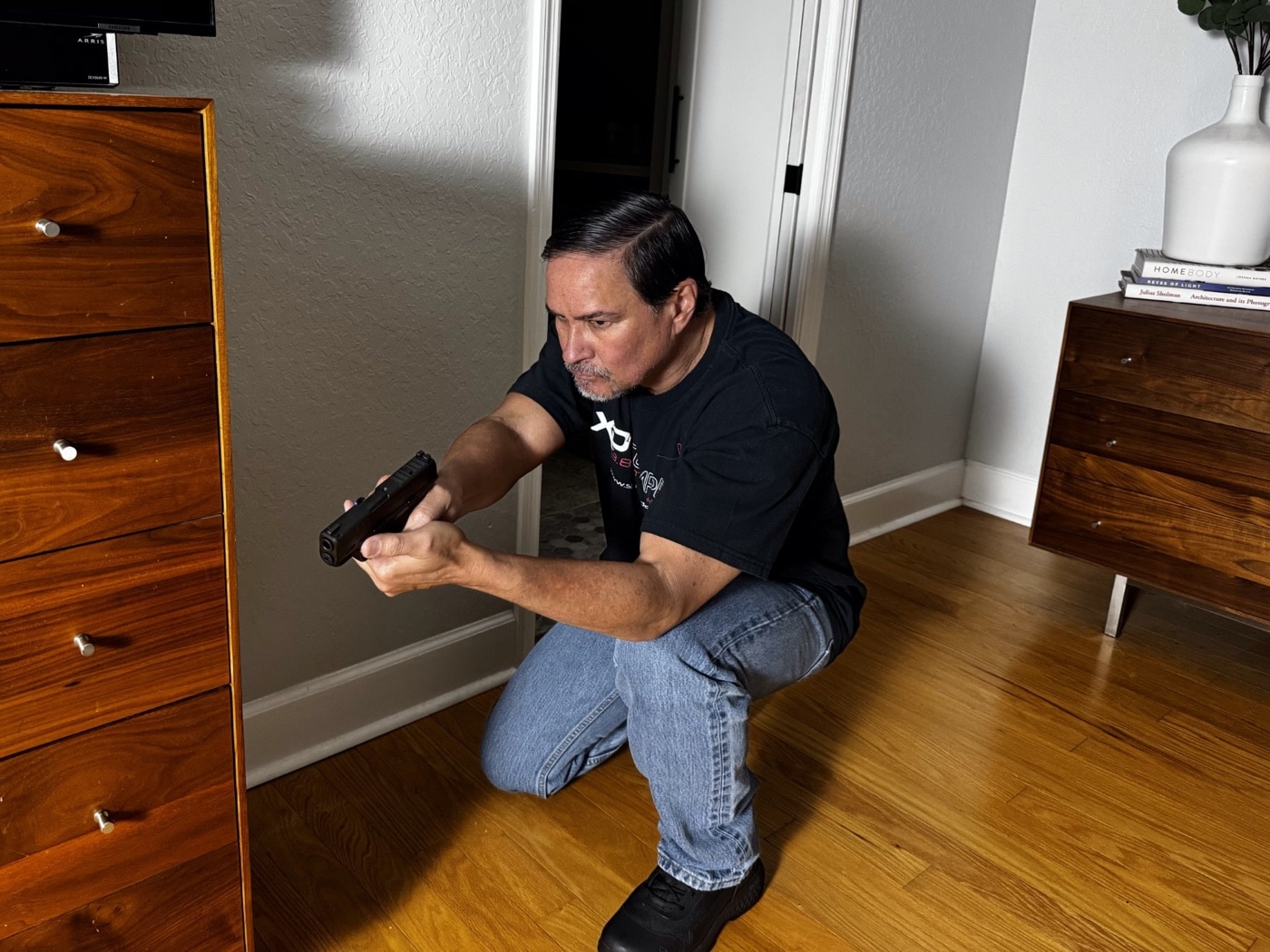 The author is pictured aiming his Springfield Hellcat handgun while crouched behind a large, sturdy piece of furniture, demonstrating a defensive position during a home invasion scenario. This image shows the importance of choosing cover instead of just concealment when facing an intruder. Heavy furniture, such as a couch or solid table, provides better protection from gunfire or physical attacks compared to lighter or thinner objects. The person keeps the firearm ready but maintains a low profile to minimize exposure. Learning how to survive a home invasion involves using available cover, knowing your home’s layout, and staying calm under pressure. The use of a defensive firearm like the Springfield Hellcat is also part of personal home safety strategies. Practicing these defensive tactics can increase your chances of surviving an encounter with a violent intruder.