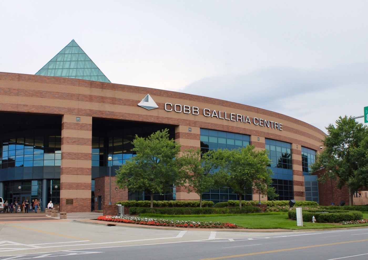 This image shows the exterior of the Cobb Galleria Centre in Atlanta, Georgia, taken during the 2025 Blade Show. The front facade features large glass windows framed by brick and metal elements under a clear sky. Blade Show signage and promotional banners are visible near the entrance, indicating the event’s presence. The wide plaza in front of the building is empty of people but clearly set up for a crowd, with planned walkways and entry points. Modern architectural details, like a pyramid skylight and overhanging roofs, stand out. The photo communicates a professional conference setting about to welcome thousands of attendees.