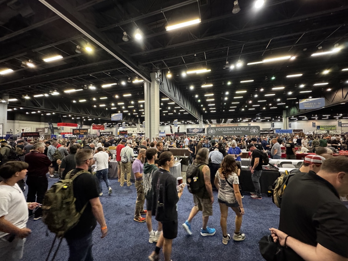 This image shows a group of Blade Show 2025 attendees standing and walking throughout the main convention hall. The crowd is diverse, including people of various ages and backgrounds, many wearing lanyards or event badges. Some attendees are carrying tote bags, backpacks, or notebooks as they move between booths. The mood is lively, with groups stopping to chat, take photos, or discuss products with exhibitors. Bright overhead lighting fills the hall, and there are visible banners and booth signs in the background. The scene captures the social and community aspect of the Blade Show, highlighting how the event brings together knife enthusiasts and industry professionals.