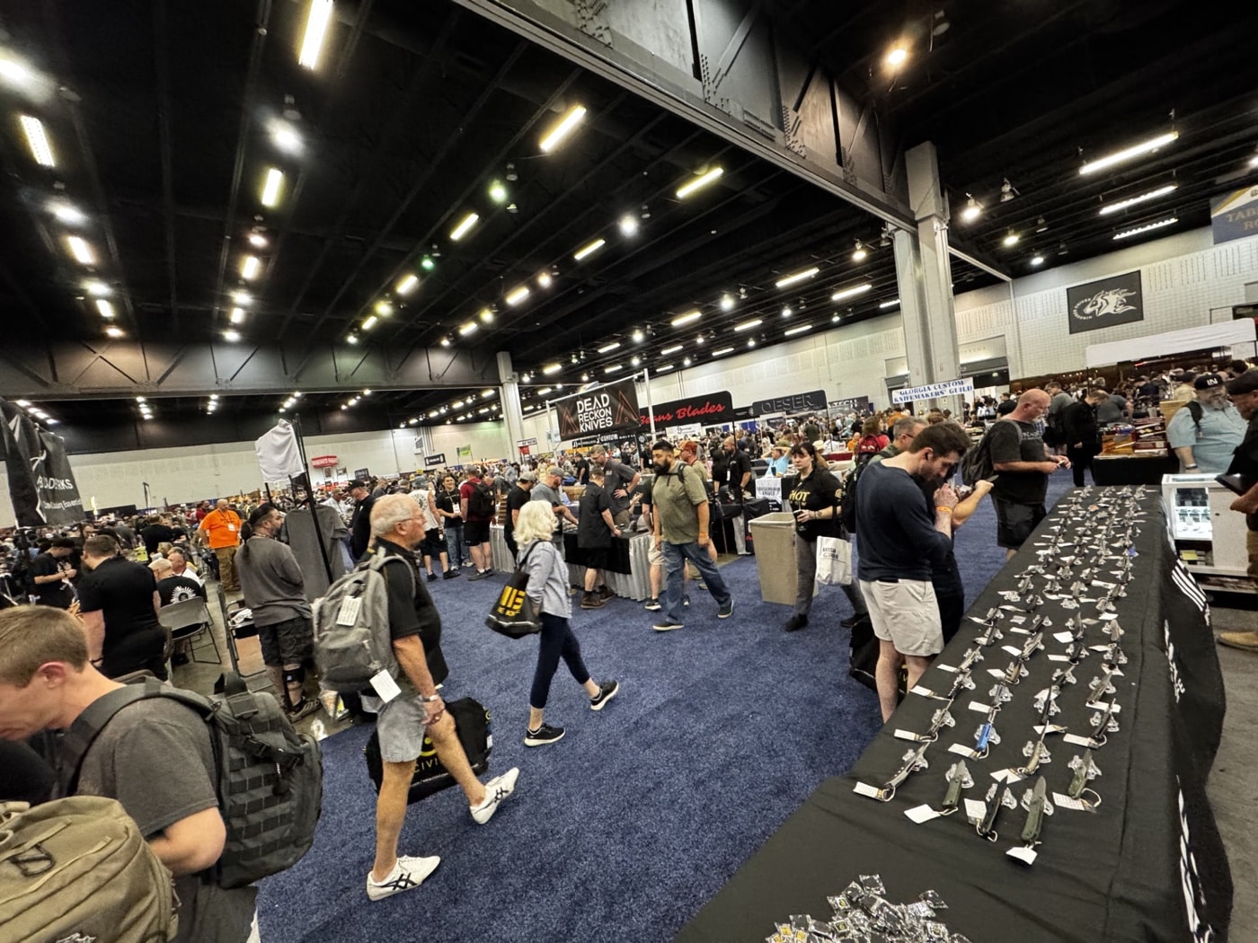 This image captures a wide view of the 2025 Blade Show floor at the Cobb Galleria in Atlanta. Rows of exhibitor booths line both sides of the main aisle, each showcasing knives, tools, and accessories. Dozens of attendees move through the aisles, some stopping to handle knives or talk with booth representatives, while others walk and browse. Booth banners, branded displays, and informational signs are visible throughout the scene. The crowd is a mix of collectors, makers, and industry professionals, many wearing lanyards or event badges. The lighting is bright and even, emphasizing the scale and busy atmosphere of the world’s largest knife show. This image highlights the hands-on, interactive nature of the event.