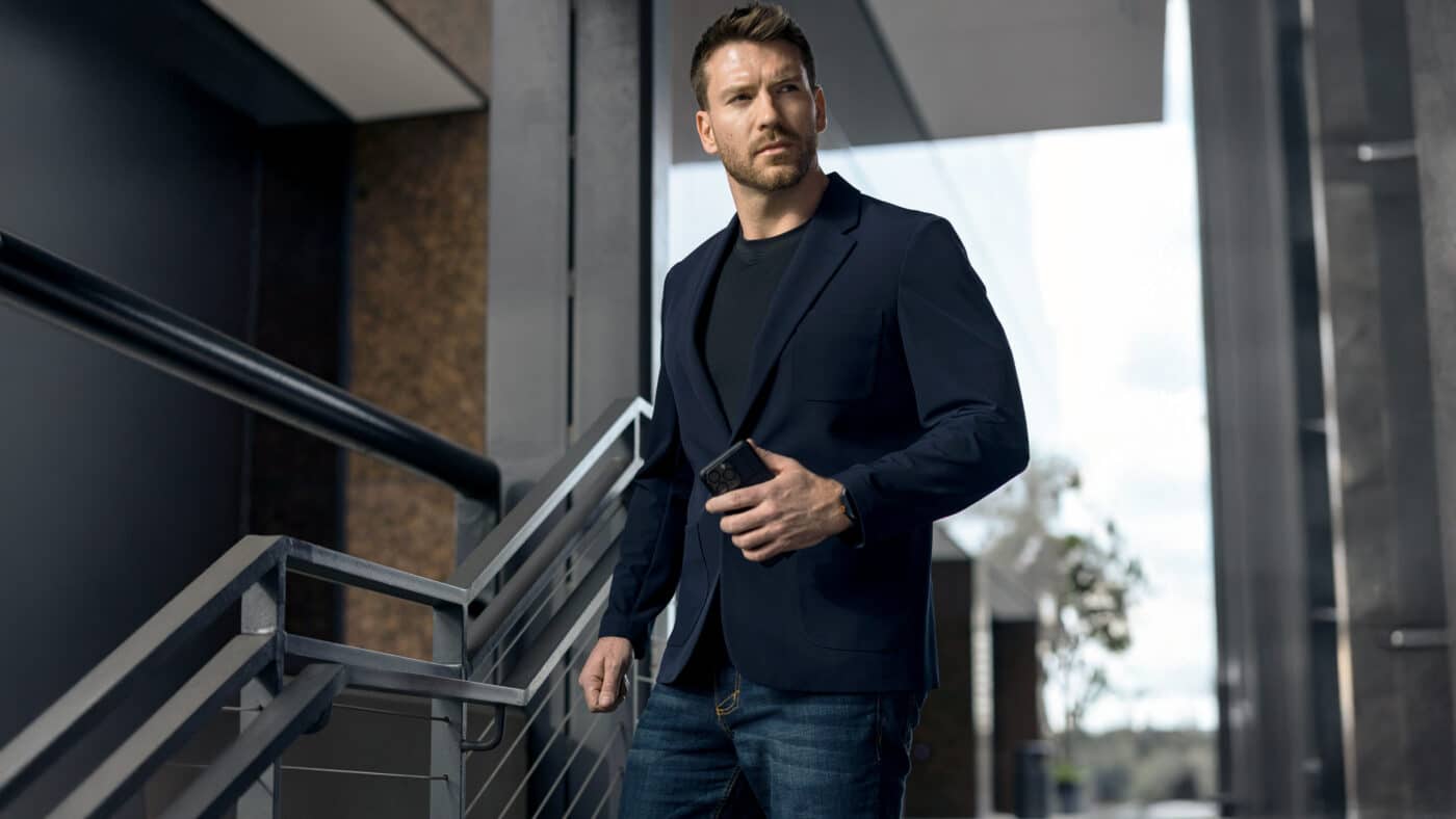 This image shows the 5.11 Founder’s Jacket displayed upright on a mannequin, with the jacket fully buttoned and visible from the front. The fabric has a smooth, matte finish typical of a sport jacket, in dark navy or black. The jacket’s clean lines and professional cut make it look like standard business-casual outerwear, but it’s built with tactical features for concealed carry and gear storage. There are no visible logos or military-style elements, helping it blend in for low-profile security or civilian use. Subtle details like reinforced stitching and discreet pocket seams hint at extra durability and utility. This product photo is meant for a 5.11 Founder’s Jacket review article, focusing on both appearance and function. The overall presentation shows a jacket suitable for both armed security work and daily wear.