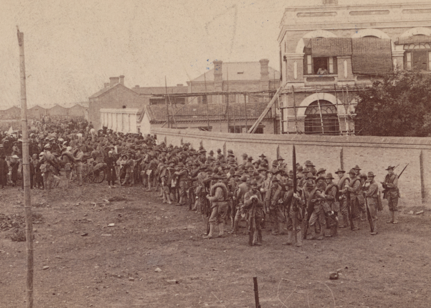 The photograph shows U.S. 9th Infantry soldiers gathered in a large field on the outskirts of Tientsin (Tianjin), China, preparing to begin their march to relieve the Siege of the International Legations in Peking. Troops are dressed in campaign uniforms with rifles slung and full field packs, standing in loose formation or resting on the ground while officers organize final preparations. Piles of supplies, ammunition crates, and horses or wagons are visible nearby, underscoring the scale of the operation. The flat landscape stretches out behind the men, with the city of Tientsin’s rooftops or distant walls visible in the background. Some soldiers confer quietly while others check equipment or fill canteens. The scene captures the tense anticipation before a major advance, showing the logistical coordination and readiness of American forces. The photo provides historical context for the start of the relief mission that would help end the 55-day siege in Peking.
