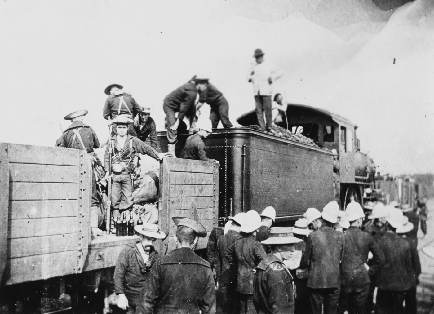 The photograph shows a busy scene at a railway station in northern China as Allied troops — including Americans, British, Japanese, Russians, and other nationalities — board a train bound for Peking during the Boxer Rebellion. Soldiers in a variety of uniforms, some with rifles slung and heavy packs on their backs, crowd the platform and climb into open railcars or carriages. Officers direct the boarding process while porters or local workers load supplies, ammunition, and food onto the train. The train itself consists of steam locomotives and boxcars or open flatcars modified for troop transport. The atmosphere is tense but determined, with men preparing for the dangerous advance to relieve the besieged Legation Quarter. The background includes telegraph poles, stacks of crates, and the broad expanse of Chinese countryside or a makeshift station. The photo captures the multinational nature of the relief force and the logistical effort required to move troops quickly toward the front lines.