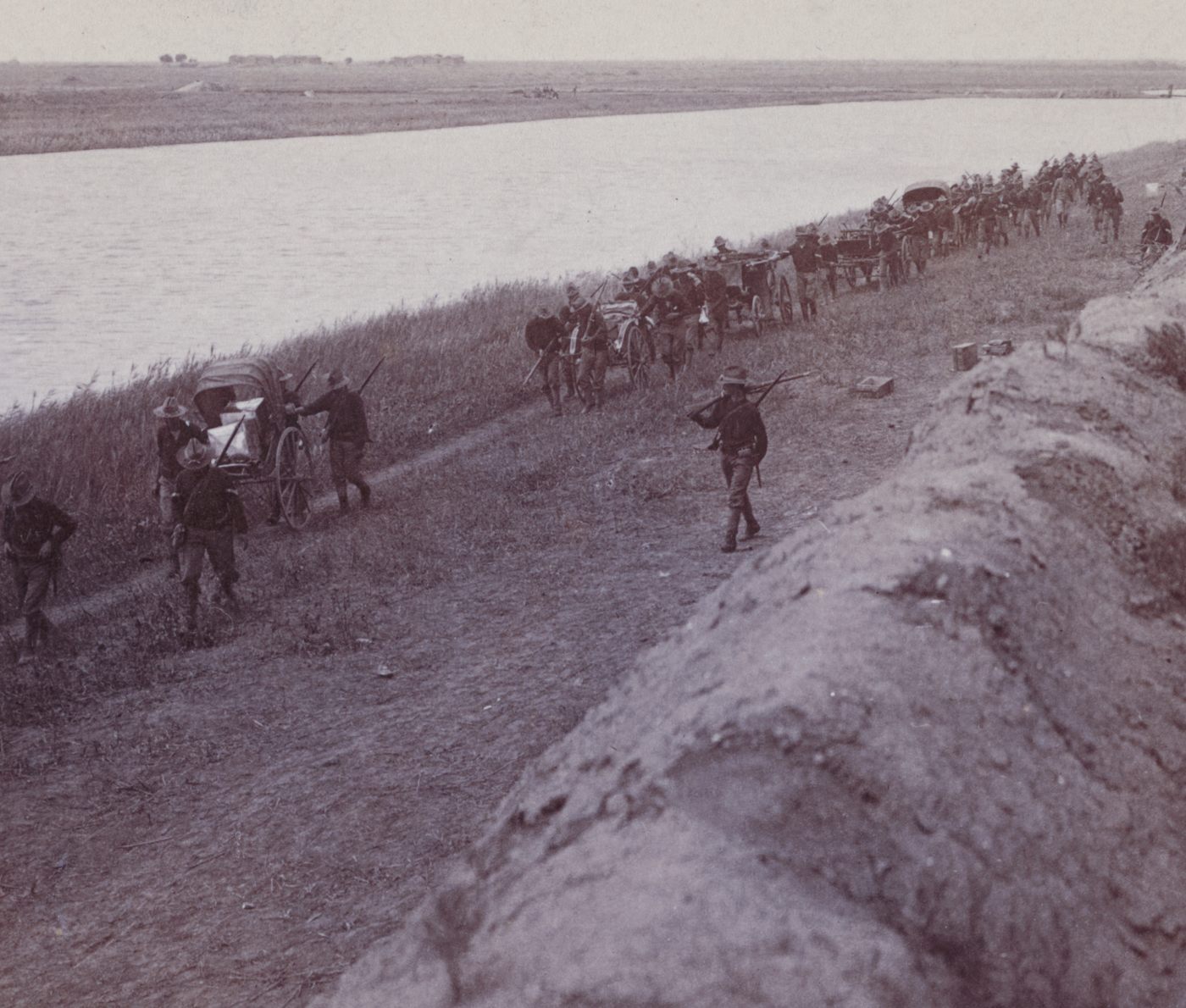 The photograph shows a group of American troops — most likely infantry or Marines — positioned near a large mud wall in Tientsin (Tianjin), China, during the Boxer Rebellion. The soldiers are in field uniforms with campaign hats, carrying rifles and packs, and are seen either crouching behind the wall for cover or moving in formation alongside it. The mud wall is high and thick, made of packed earth, with an uneven, rough surface and occasional cracks or damage from recent fighting. Some soldiers scan the area over the wall, while others converse, tend to equipment, or stand guard. The surroundings include patches of bare ground, scattered debris, and possibly local buildings or other defensive structures in the background. The image reflects the gritty urban combat and the use of improvised fortifications in the battle for Tientsin. It highlights the challenges American forces faced in navigating and fighting around such barriers during the China Relief Expedition.