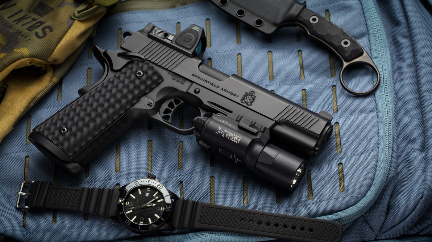 The photo displays a Springfield Armory 1911 pistol positioned beside an ArmourLite watch on a neutral background. The watch’s robust build and illuminated dial are visible, suggesting its use in tactical or demanding environments. The pistol is shown with clean lines and recognizable branding, reinforcing its reputation for reliability and precision. This image is used to visually connect the interview article’s themes of toughness, performance, and attention to detail. Both items are placed to highlight their features without overlap, making it clear each represents a different aspect of professional gear. The photo supports the article’s focus on the intersection between high-quality firearms and advanced watchmaking. It’s relevant for readers interested in gear built for military, law enforcement, and outdoor use.